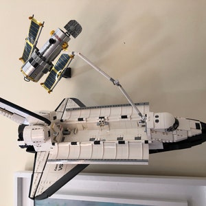 Puede incluir: Una lanzadera espacial Lego blanca y negra con un brazo rob&oacute;tico que se extiende hacia una estaci&oacute;n espacial Lego plateada y dorada. La lanzadera espacial tiene el texto "Discovery" y "STS" en el lateral.