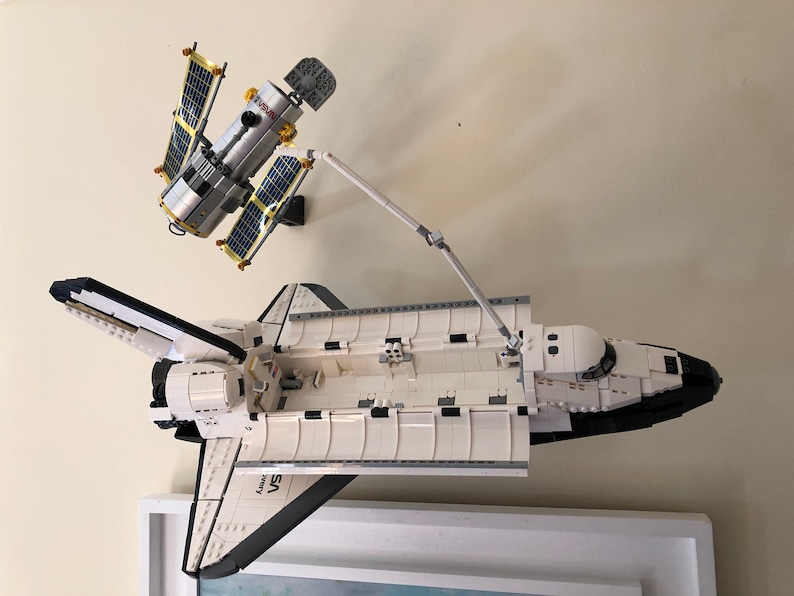 Puede incluir: Una lanzadera espacial Lego blanca y negra con un brazo rob&oacute;tico que se extiende hacia una estaci&oacute;n espacial Lego plateada y dorada. La lanzadera espacial tiene el texto "Discovery" y "STS" en el lateral.