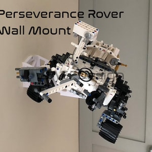 May include: A white Lego model of the Perseverance Rover, mounted on a wall. The model is detailed and includes the rover's wheels, arms, and cameras. The text "Perseverance Rover Wall Mount" is visible in the image.