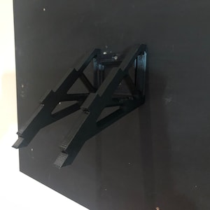 May include: Black metal wall mount for a laptop or tablet. The mount is designed with a triangular shape and has a minimalist aesthetic.
