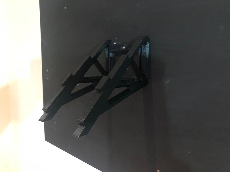 May include: Black metal wall mount for a laptop or tablet. The mount is designed with a triangular shape and has a minimalist aesthetic.