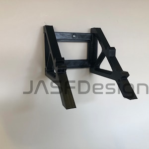 May include: A black 3D printed wall mount for a laptop or tablet. The mount has a simple design with two angled supports and a rectangular cutout in the center. The text "JASFDesign" is visible in the lower center of the image.