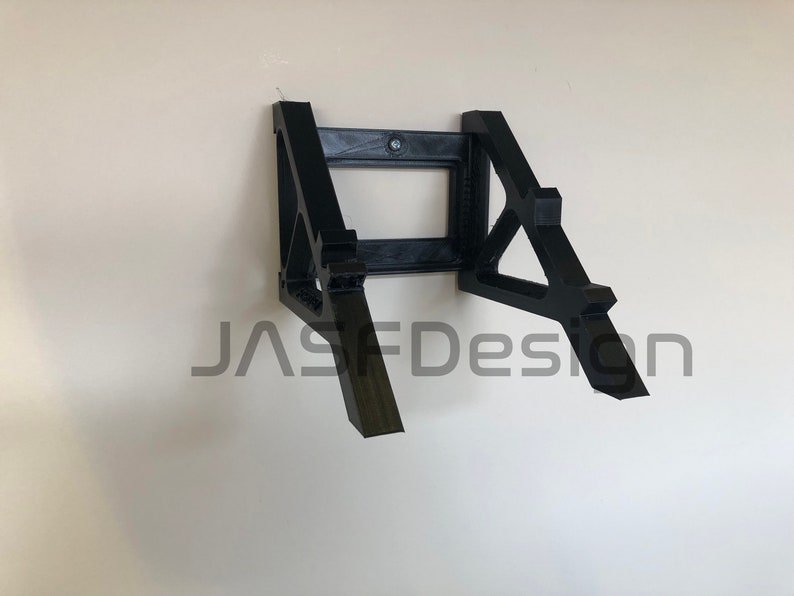 May include: A black 3D printed wall mount for a laptop or tablet. The mount has a simple design with two angled supports and a rectangular cutout in the center. The text "JASFDesign" is visible in the lower center of the image.