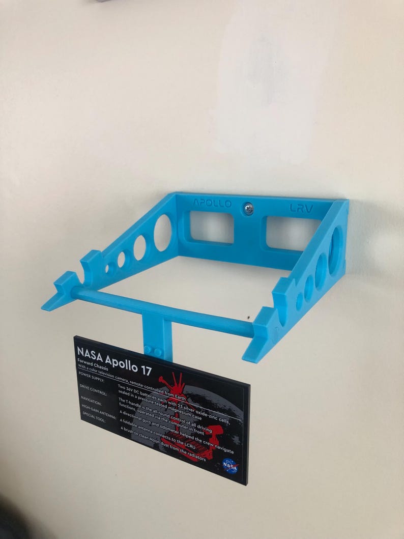 May include: A blue 3D printed model of the Lunar Roving Vehicle (LRV) used by NASA during the Apollo 17 mission. The model is mounted on a wall and includes a small black plaque with information about the LRV.