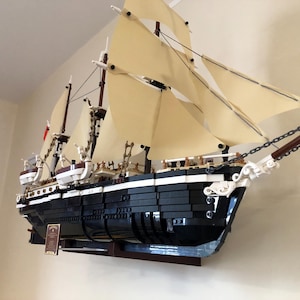May include: A black and white LEGO model of a ship with white sails, mounted on a wooden wall shelf. The ship has a detailed hull and rigging, and is a popular collectible for LEGO enthusiasts.
