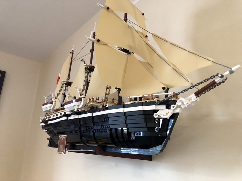May include: A black and white LEGO model of a ship with white sails, mounted on a wooden wall shelf. The ship has a detailed hull and rigging, and is a popular collectible for LEGO enthusiasts.