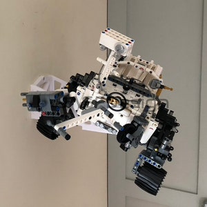 May include: A white and gray Lego model of the Mars rover Curiosity. The rover has six wheels, a robotic arm, and a camera. The model is detailed and realistic.