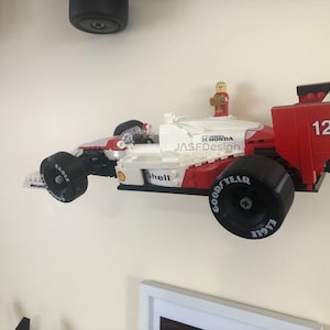 Op de afbeelding: Een rood, wit en zwart Lego raceauto model met nummer 12 aan de zijkant. De auto heeft Goodyear Eagle banden en Shell branding. Een Lego figuur staat bovenop de auto. De auto is aan een witte muur bevestigd.