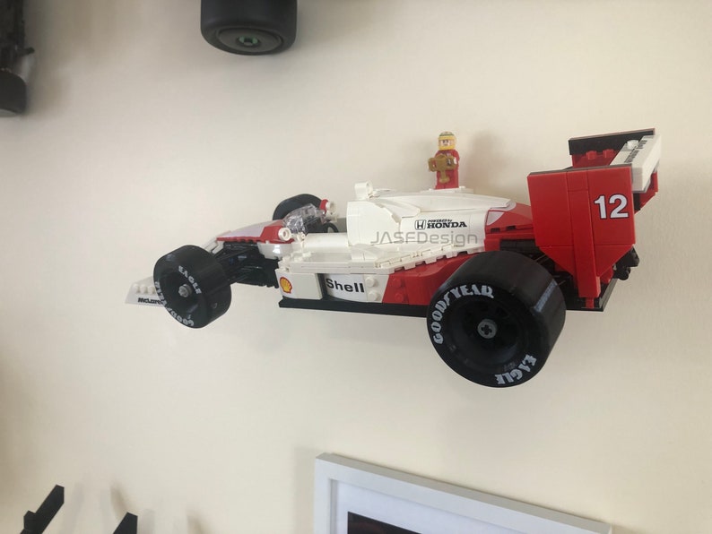 Op de afbeelding: Een rood, wit en zwart Lego raceauto model met nummer 12 aan de zijkant. De auto heeft Goodyear Eagle banden en Shell branding. Een Lego figuur staat bovenop de auto. De auto is aan een witte muur bevestigd.