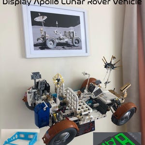 May include: A 3D printed wall mount for displaying a model of the Apollo Lunar Rover. The mount is made of white plastic and has a blue base. The model is made of white, brown, and blue plastic and has a detailed design. The image also includes a framed photo of the Apollo Lunar Rover on the moon. The text "STL Files Not a Lego Product" is visible in the image.