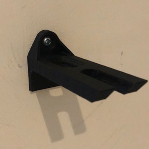 May include: A black plastic wall mount with a single screw. The mount has two slots for holding objects.