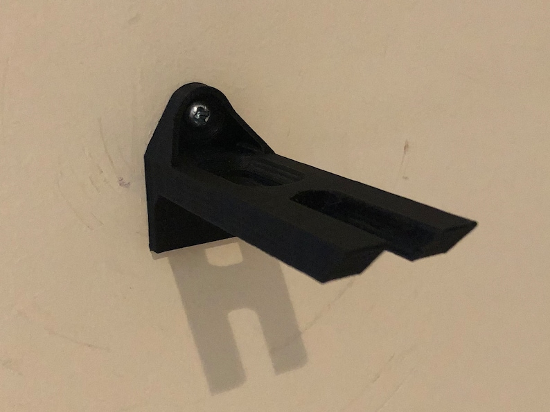 May include: A black plastic wall mount with a single screw. The mount has two slots for holding objects.