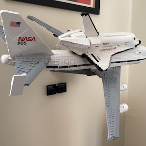 May include: A white and gray LEGO model of a NASA space shuttle atop a modified Boeing 747. The model features the NASA logo, American flag, and the number 905. The model is mounted on a wall.