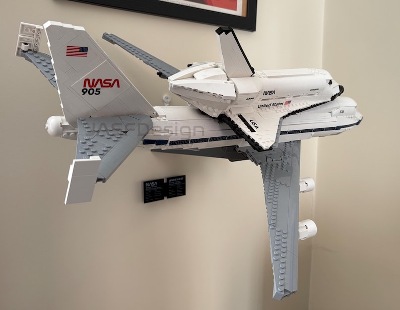 May include: A white and gray LEGO model of a NASA space shuttle atop a modified Boeing 747. The model features the NASA logo, American flag, and the number 905. The model is mounted on a wall.