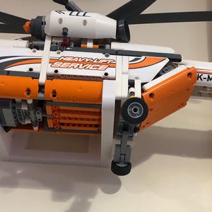 May include: A white and orange Lego helicopter model with the text "Heavy-Lift Service" and "K-MLX" on the side. The helicopter is mounted on a white stand.
