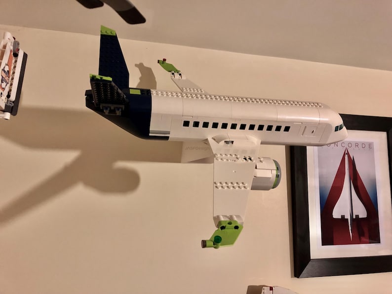 May include: A white, green, and blue Lego airplane model mounted on a wall. The airplane has a white fuselage, green wings, and a blue tail. The model is mounted on a wall with a black frame around it. The frame has the word "CONCORDE" printed on it.