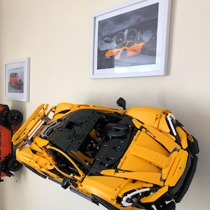 May include: A large, yellow and black Lego model of a sports car is mounted on a wall. The model is highly detailed, with visible engine components and aerodynamic features. Two framed pictures are also on the wall, one with a red car and the other with an orange sports car.