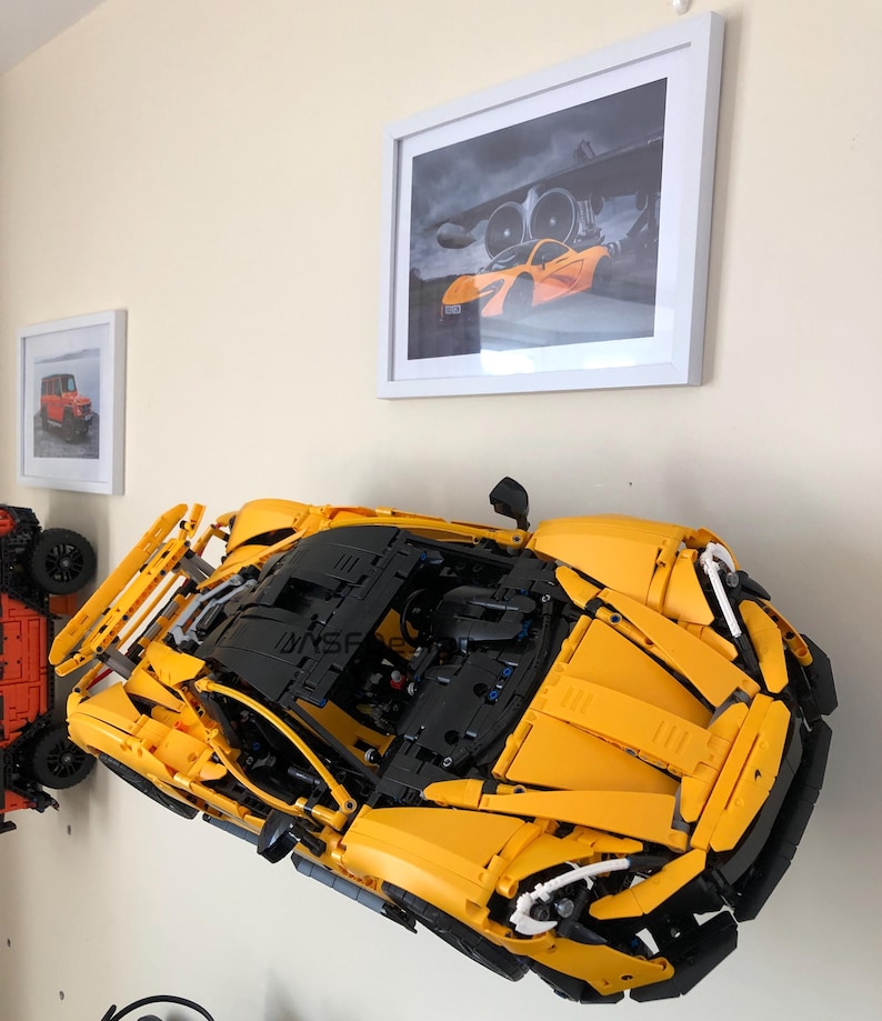 May include: A large, yellow and black Lego model of a sports car is mounted on a wall. The model is highly detailed, with visible engine components and aerodynamic features. Two framed pictures are also on the wall, one with a red car and the other with an orange sports car.