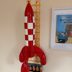 Puede incluir: Un modelo de cohete rojo y blanco con una estructura de soporte amarilla, montado en la pared. El cohete tiene un patr&oacute;n de cuadros y est&aacute; acompa&ntilde;ado de figuras de astronautas en miniatura. Un p&oacute;ster enmarcado de "Destination Moon" es visible.