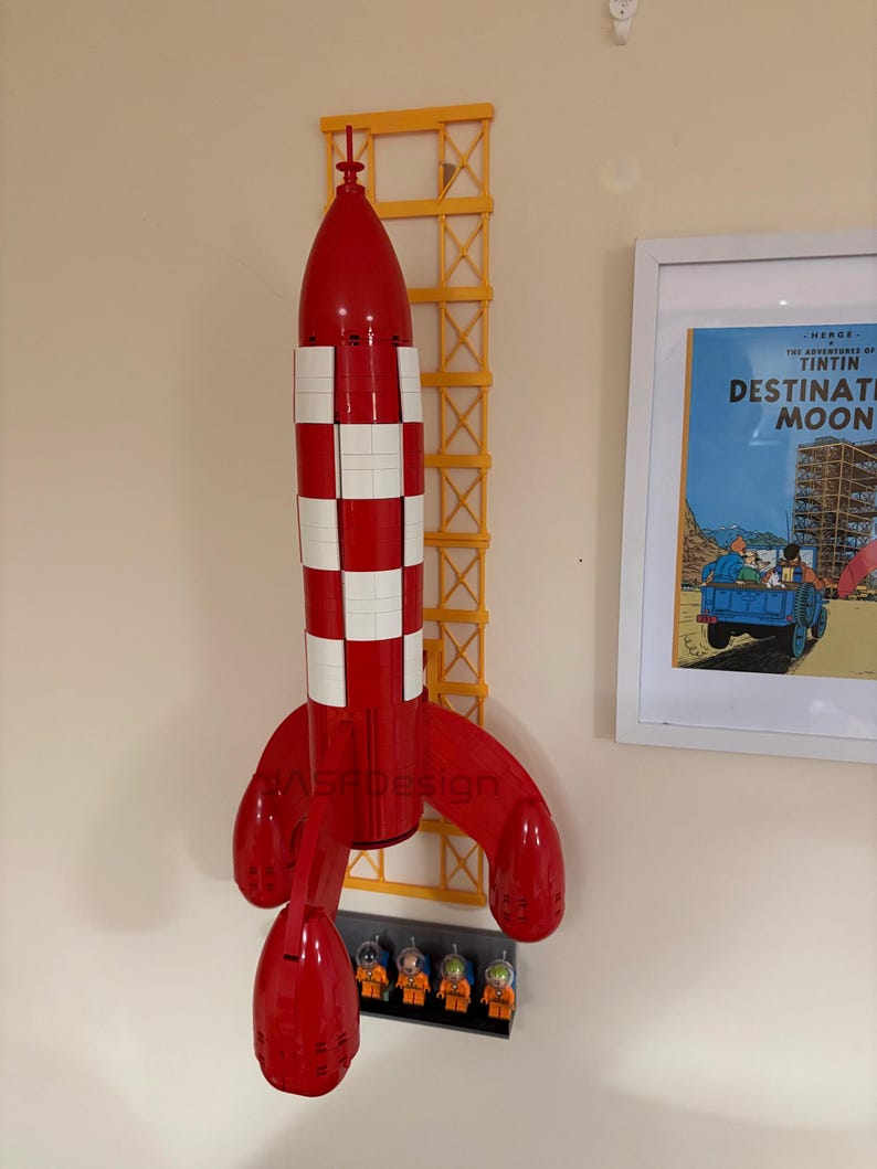 Puede incluir: Un modelo de cohete rojo y blanco con una estructura de soporte amarilla, montado en la pared. El cohete tiene un patr&oacute;n de cuadros y est&aacute; acompa&ntilde;ado de figuras de astronautas en miniatura. Un p&oacute;ster enmarcado de "Destination Moon" es visible.
