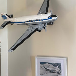 May include: A white and gray model airplane with the words "PAN AMERICAN" printed on the side. The model is mounted on a wall, with a framed black and white photograph of a similar airplane below it.