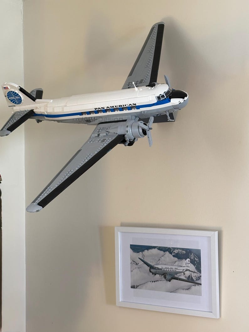 May include: A white and gray model airplane with the words "PAN AMERICAN" printed on the side. The model is mounted on a wall, with a framed black and white photograph of a similar airplane below it.
