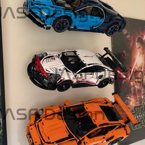 May include: Three Lego car models displayed on a wall. The top car is blue and black, the middle car is white and red, and the bottom car is orange.