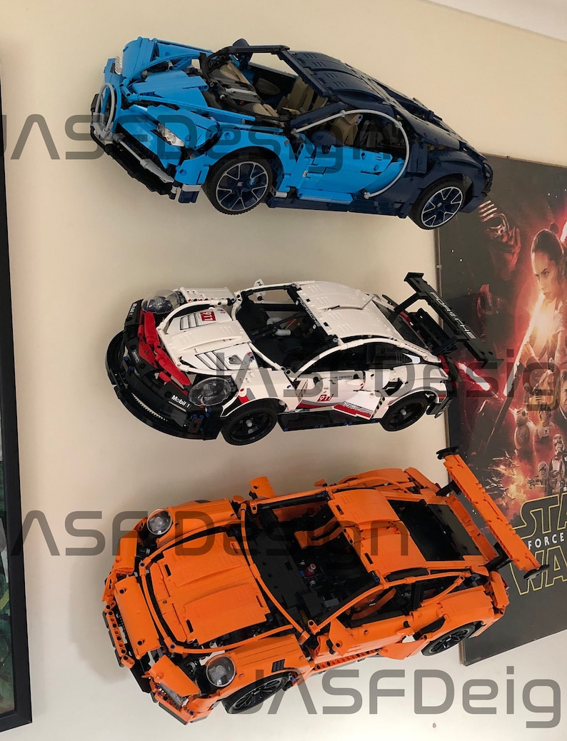 May include: Three Lego car models displayed on a wall. The top car is blue and black, the middle car is white and red, and the bottom car is orange.