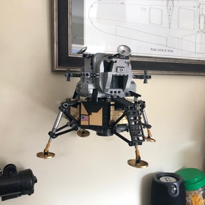 May include: A silver and black Lego model of the lunar module, the spacecraft that landed on the moon. The model has a gold-colored American flag on the side and is mounted on a wall.