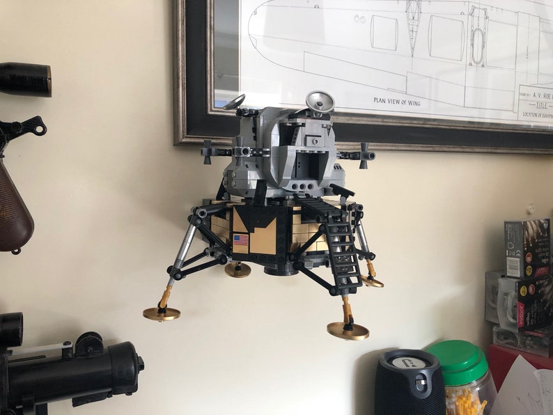 May include: A silver and black Lego model of the lunar module, the spacecraft that landed on the moon. The model has a gold-colored American flag on the side and is mounted on a wall.