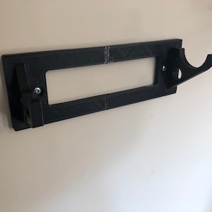 May include: A black plastic wall mount with a hook for hanging items. The mount has a rectangular opening in the center.