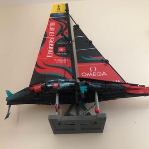 K&ouml;nnte beinhalten: Ein rotes, schwarzes und t&uuml;rkisfarbenes LEGO-Modell eines Segelboots mit den Worten "Emirates Fly Better", "Jasdfdesign", "Toyota" und "Omega" auf den Segeln. Das Modell ist auf einem grauen St&auml;nder montiert.