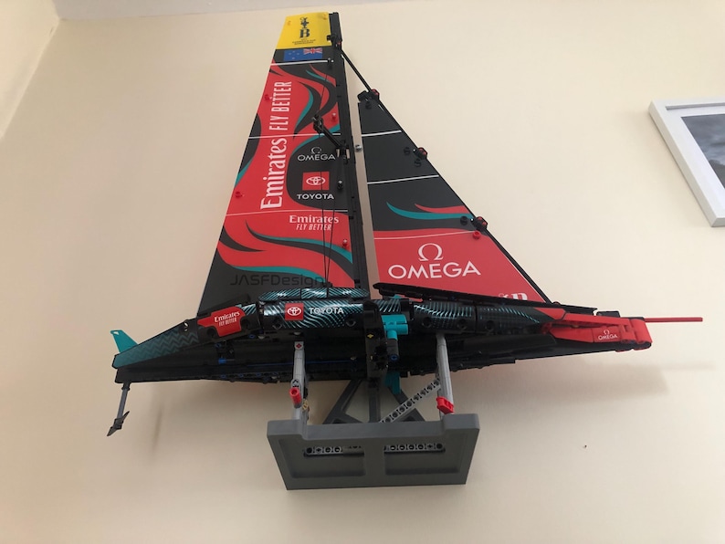 K&ouml;nnte beinhalten: Ein rotes, schwarzes und t&uuml;rkisfarbenes LEGO-Modell eines Segelboots mit den Worten "Emirates Fly Better", "Jasdfdesign", "Toyota" und "Omega" auf den Segeln. Das Modell ist auf einem grauen St&auml;nder montiert.