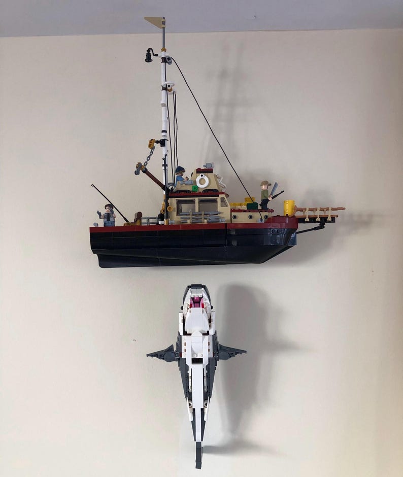 May include: A Lego model of a fishing boat with a black hull, red trim, and a white cabin. Below it is a Lego model of a shark, white with black accents, mouth open. The boat has a mast and figures.