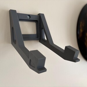 May include: A dark gray wall-mounted storage bracket featuring two parallel arms extending outward. The bracket has a rectangular base plate with a central screw hole for attachment, and each arm ends in a small upward-facing hook.