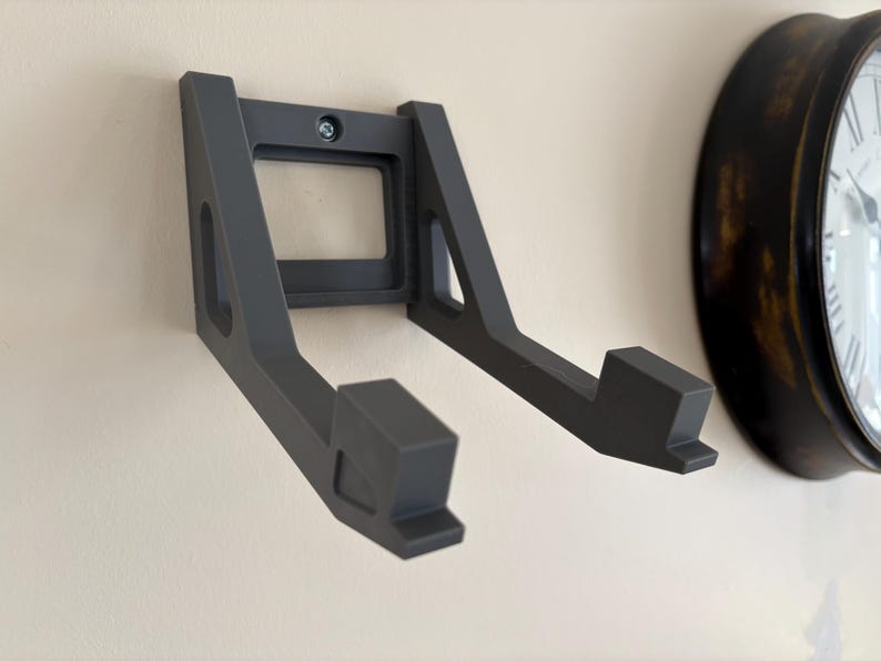 May include: A dark gray wall-mounted storage bracket featuring two parallel arms extending outward. The bracket has a rectangular base plate with a central screw hole for attachment, and each arm ends in a small upward-facing hook.