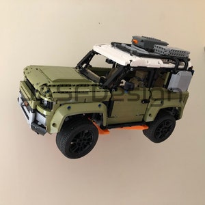 Op de afbeelding: Een groen en wit Lego Land Rover Defender model met zwarte wielen en oranje accenten. Het model is aan een muur gemonteerd.