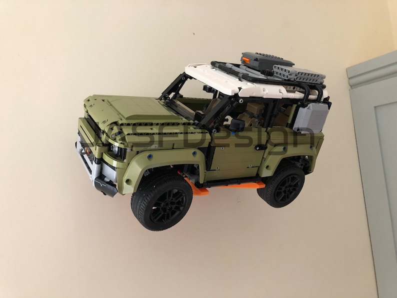 Op de afbeelding: Een groen en wit Lego Land Rover Defender model met zwarte wielen en oranje accenten. Het model is aan een muur gemonteerd.