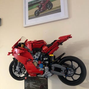 May include: A red Lego Ducati Panigale V4 R motorcycle model, built and displayed on a wall. The model is detailed with black wheels, a black exhaust, and a black seat. The model is accompanied by a black and white information card with the Ducati logo and technical specifications.