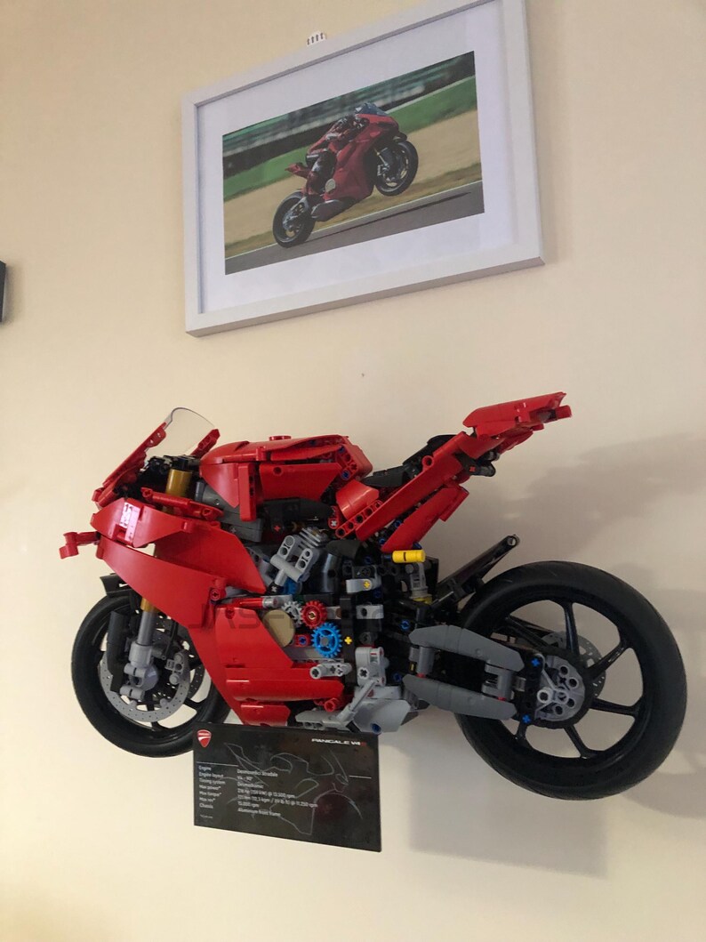 May include: A red Lego Ducati Panigale V4 R motorcycle model, built and displayed on a wall. The model is detailed with black wheels, a black exhaust, and a black seat. The model is accompanied by a black and white information card with the Ducati logo and technical specifications.