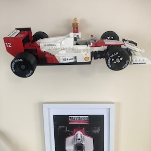 Op de afbeelding: Een rood, wit en zwart Lego-racewagenmodel met nummer 12, Goodyear-banden en Shell-branding. Het model is gemonteerd boven een ingelijste print van een vergelijkbare racewagen met Marlboro-branding.