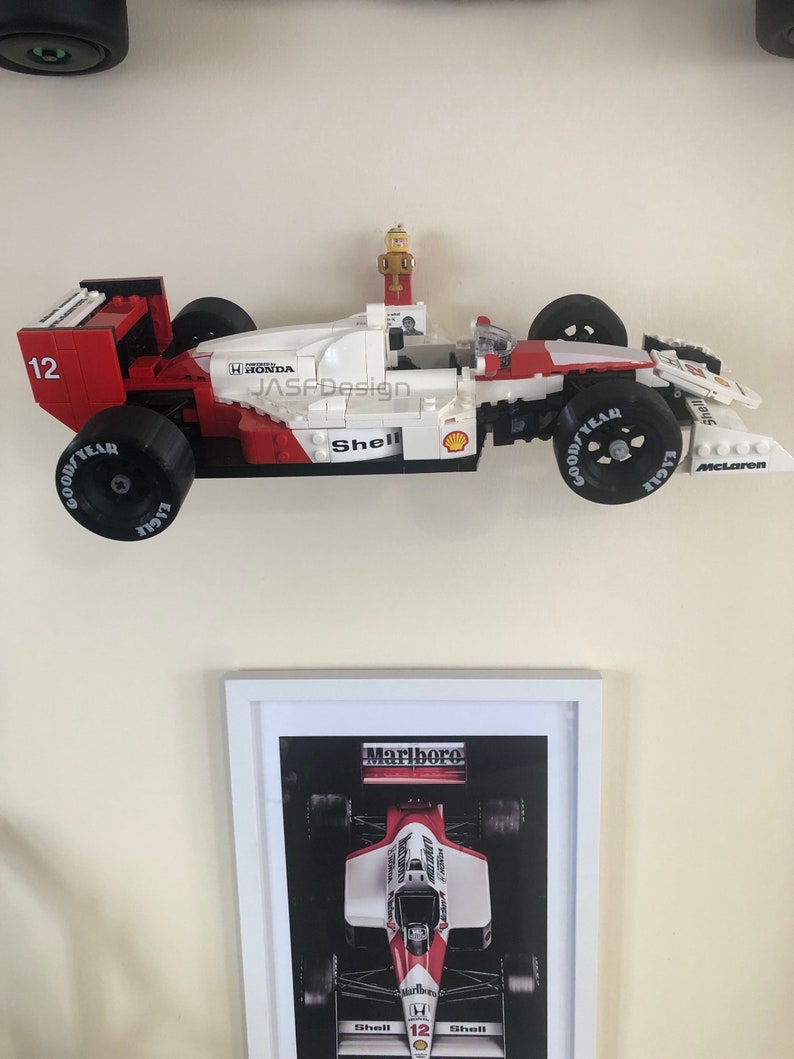 Op de afbeelding: Een rood, wit en zwart Lego-racewagenmodel met nummer 12, Goodyear-banden en Shell-branding. Het model is gemonteerd boven een ingelijste print van een vergelijkbare racewagen met Marlboro-branding.