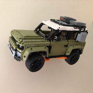 Puede incluir: Un modelo Lego de un SUV verde y blanco con ruedas negras y una baca. El coche de juguete est&aacute; montado en una pared blanca. El modelo tiene caracter&iacute;sticas detalladas.