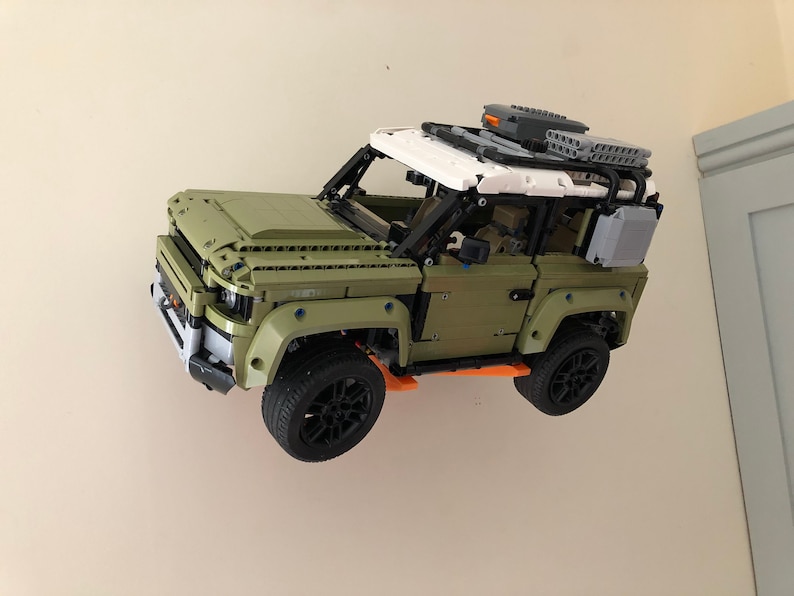 Puede incluir: Un modelo Lego de un SUV verde y blanco con ruedas negras y una baca. El coche de juguete est&aacute; montado en una pared blanca. El modelo tiene caracter&iacute;sticas detalladas.