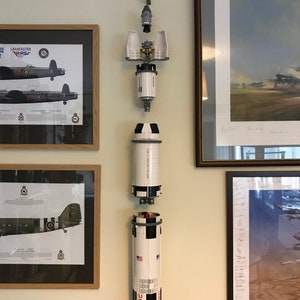 May include: A white and black Lego Saturn V rocket model is displayed on a wall, separated into its different sections. The rocket is hanging from the ceiling with string. The rocket is displayed in front of framed pictures of airplanes.