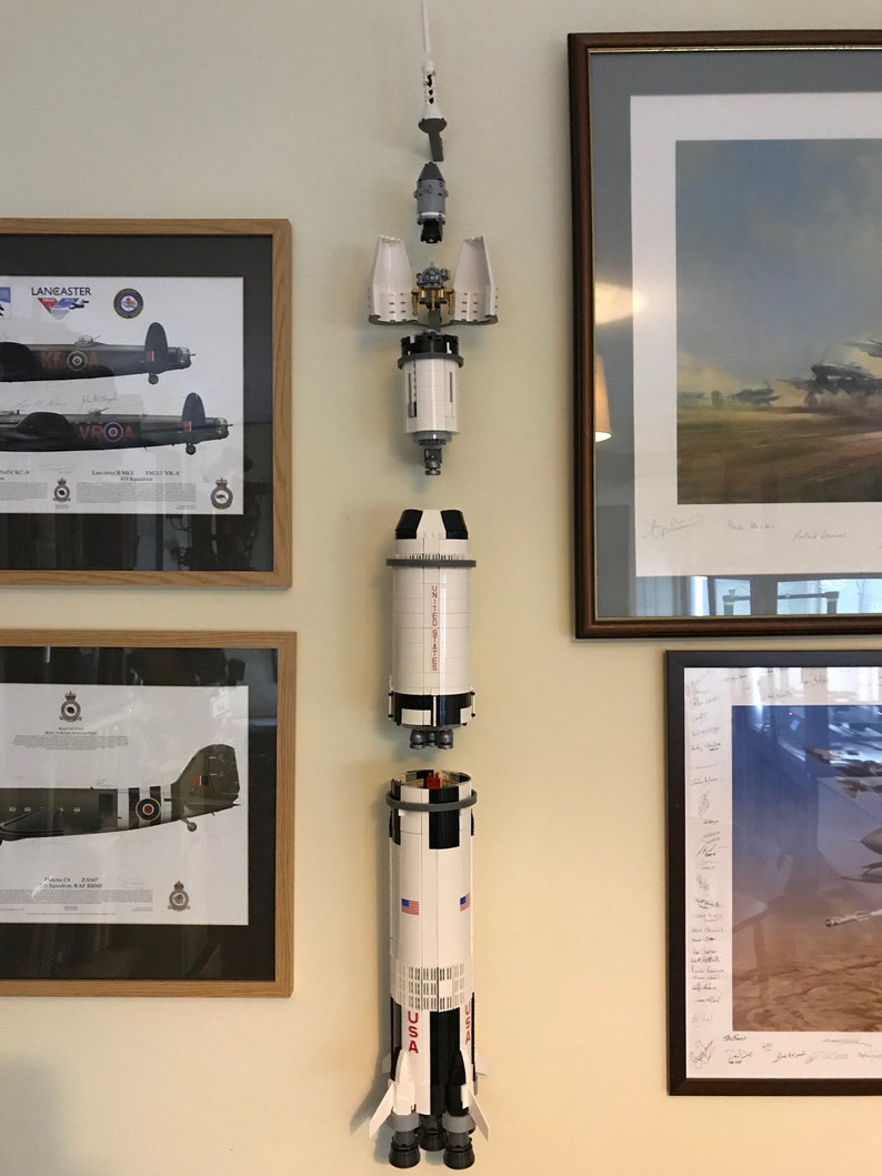 May include: A white and black Lego Saturn V rocket model is displayed on a wall, separated into its different sections. The rocket is hanging from the ceiling with string. The rocket is displayed in front of framed pictures of airplanes.