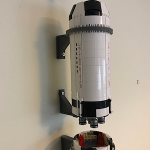 May include: A white and black Lego model of a rocket ship with the words "UNITED STATES" printed on the side. The rocket is mounted on a gray wall mount.