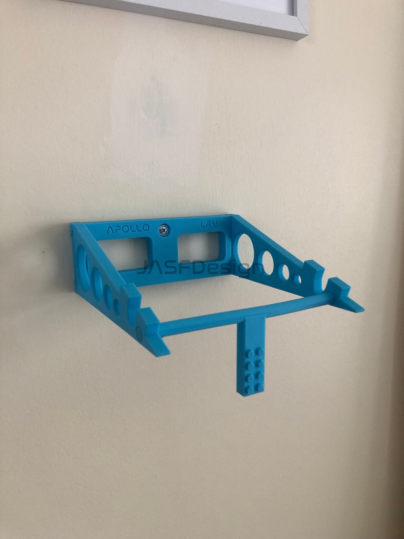 May include: A light blue, wall-mounted shelf with a geometric design. The shelf has a rectangular frame with cut-out circles and a horizontal bar. The text "APOLLO" and "CRV" are visible on the shelf. The shelf is mounted on a beige wall.