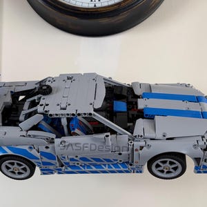 May include: A gray and blue model car made of interlocking plastic bricks. The car has blue stripes and accents, with the text "ASFDesign" visible on the side. The car's doors and hood are partially open, revealing interior details.