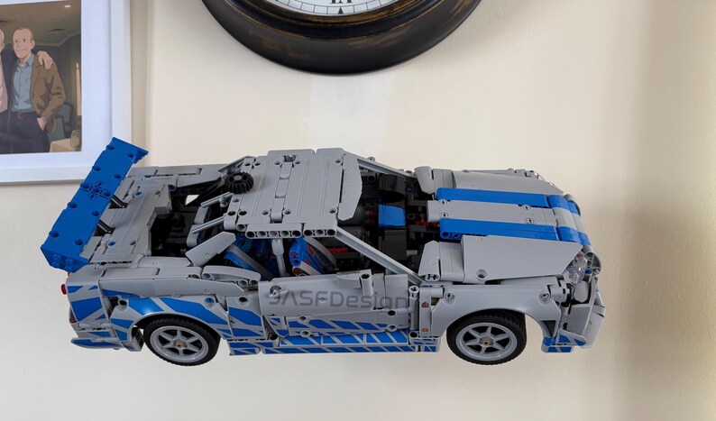 May include: A gray and blue model car made of interlocking plastic bricks. The car has blue stripes and accents, with the text "ASFDesign" visible on the side. The car's doors and hood are partially open, revealing interior details.